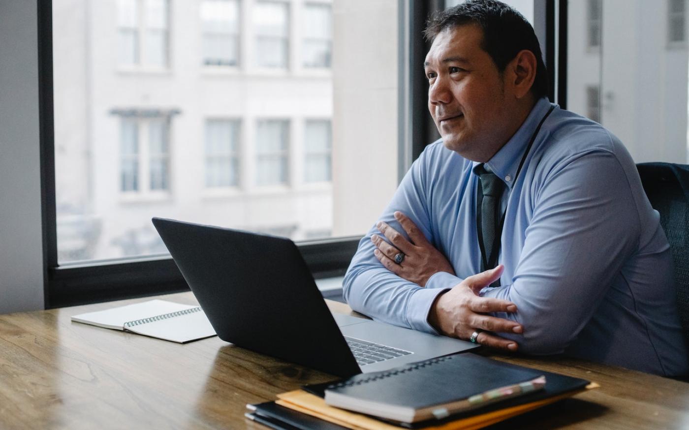 Professional-looking man sitting in an office. There is a laptop and a stack of files in front of him.