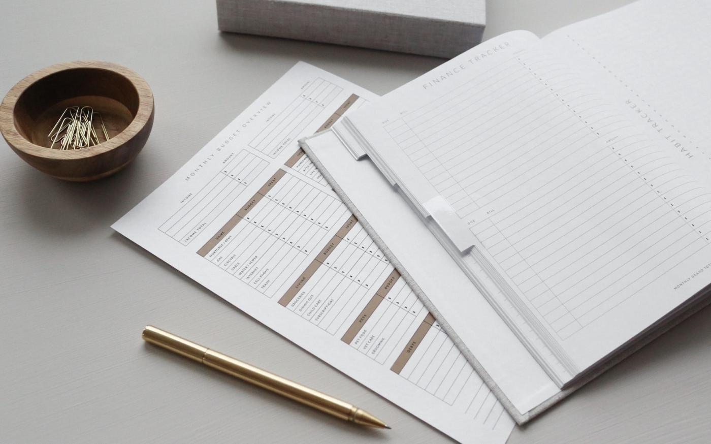 A desk with office supplies on it. In the middle of the desk is an open journal with a heading that reads "Finance Tracker". Under the journal is a piece of paper that reads "Monthly Budget Overview", and has several columns on it for tracking expenses.