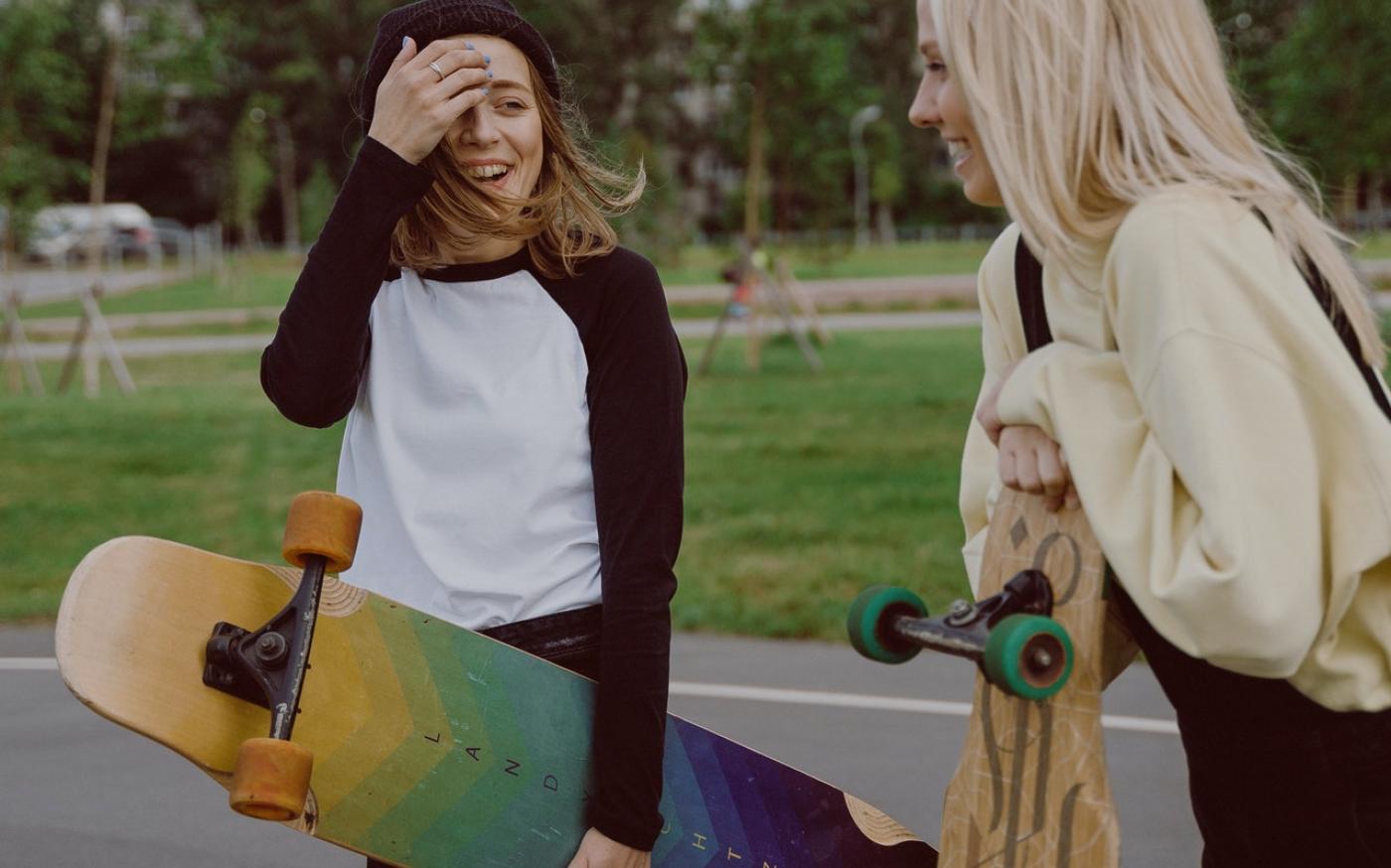 A teenager holding a yellow and blue longboard talking to her friend