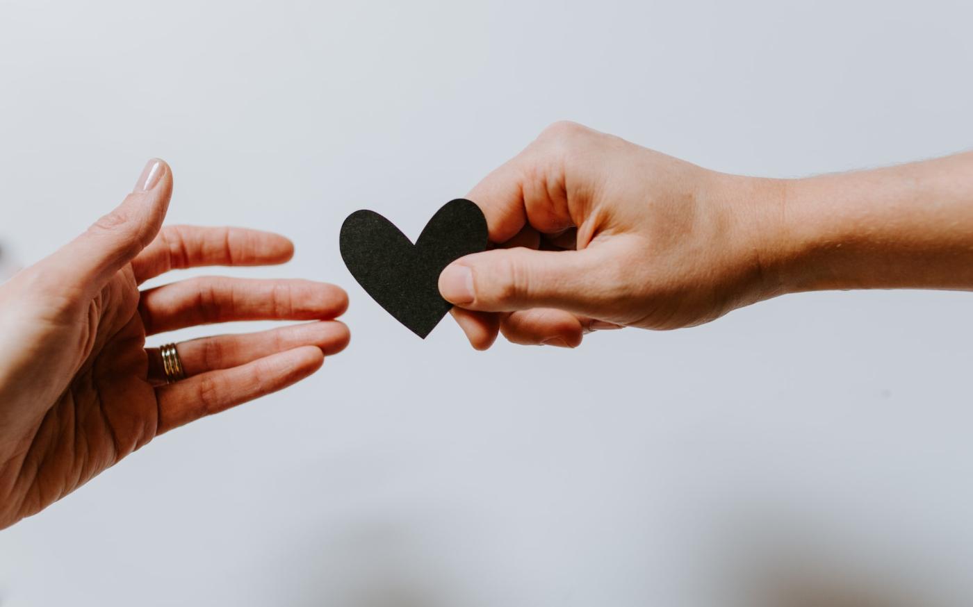 Image of two hands against a white background. One of the hands is passing a black paper heart to the other one